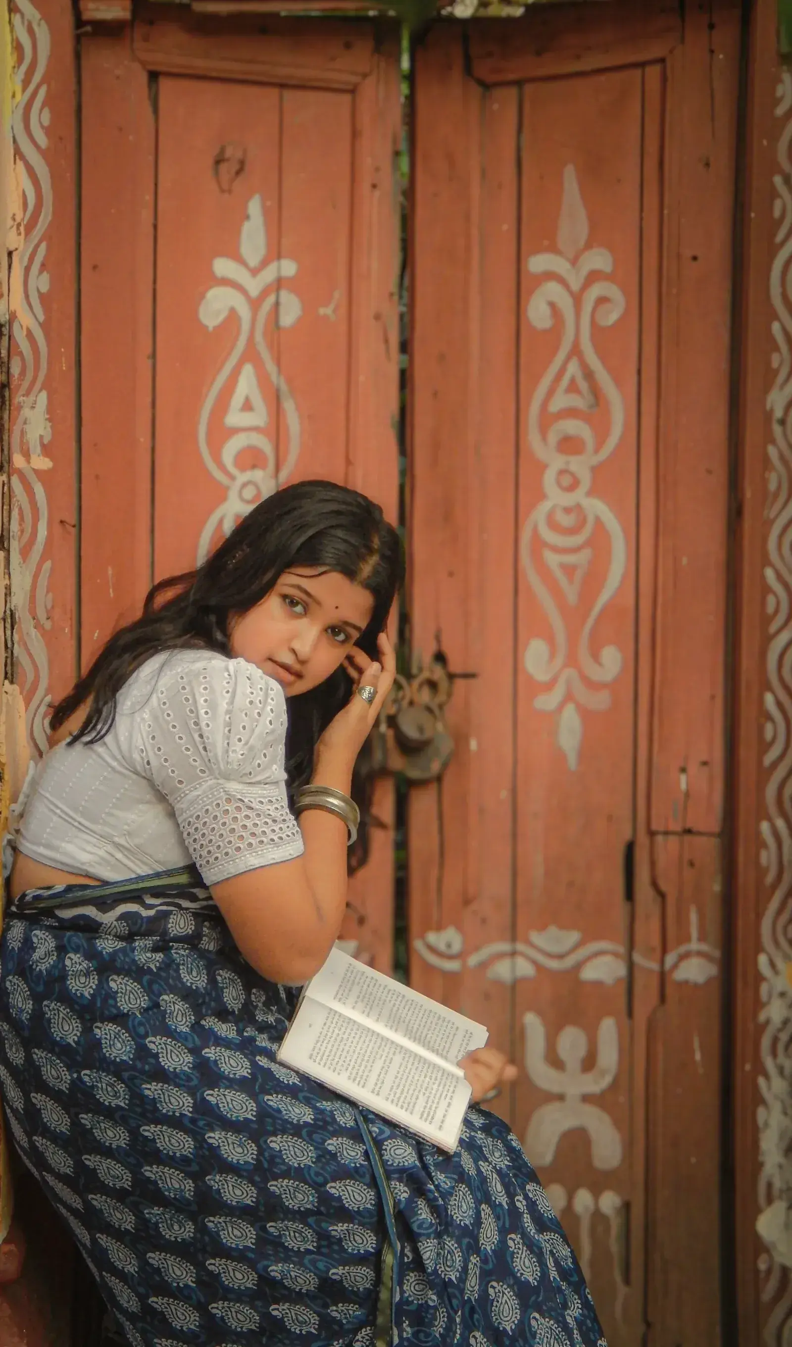 A woman in an indigo ikat saree sitting at a painted wooden door with a book
