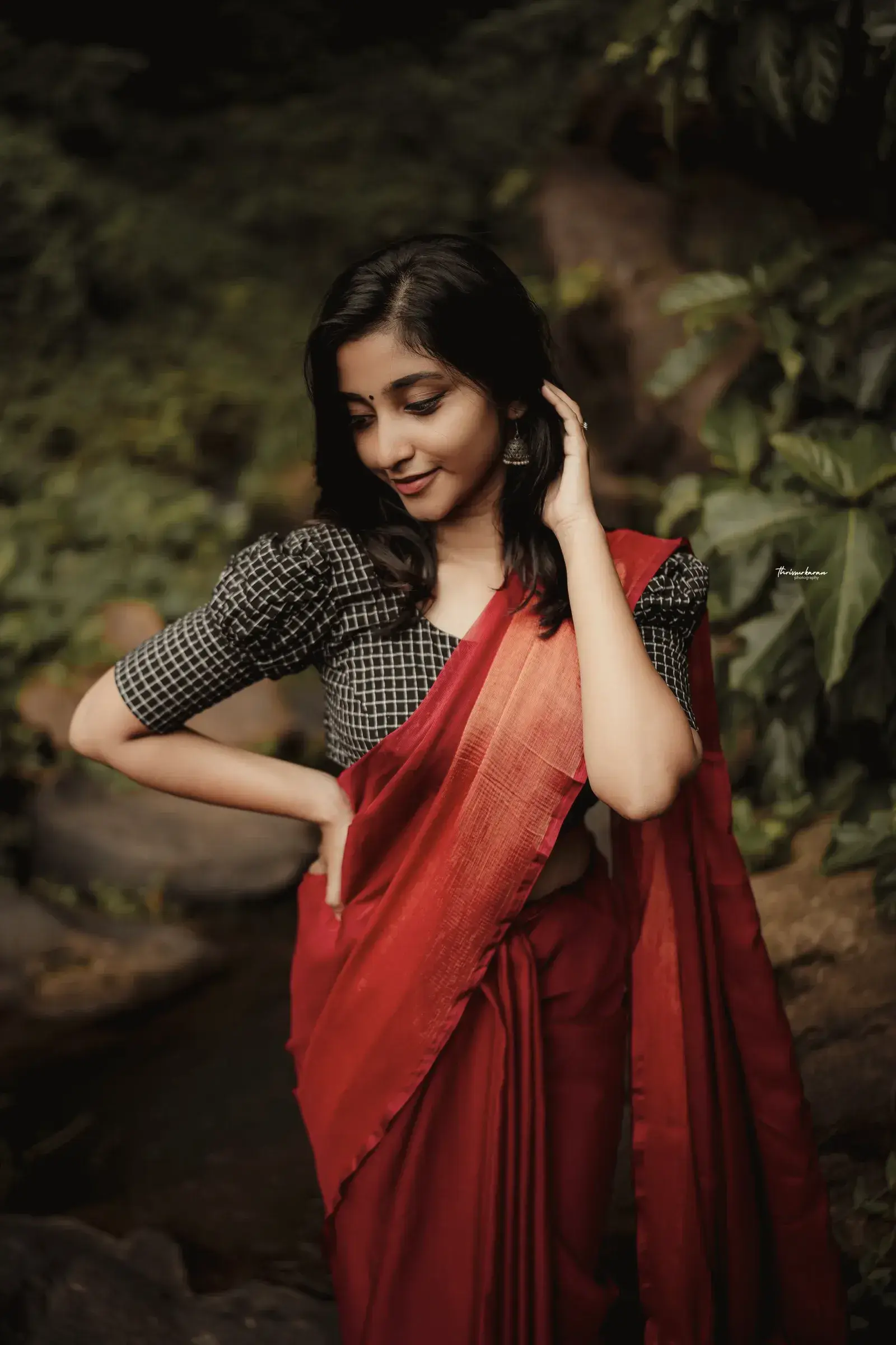 A model in a red matka silk saree and black gingham blouse, hand in her hair, among leaves