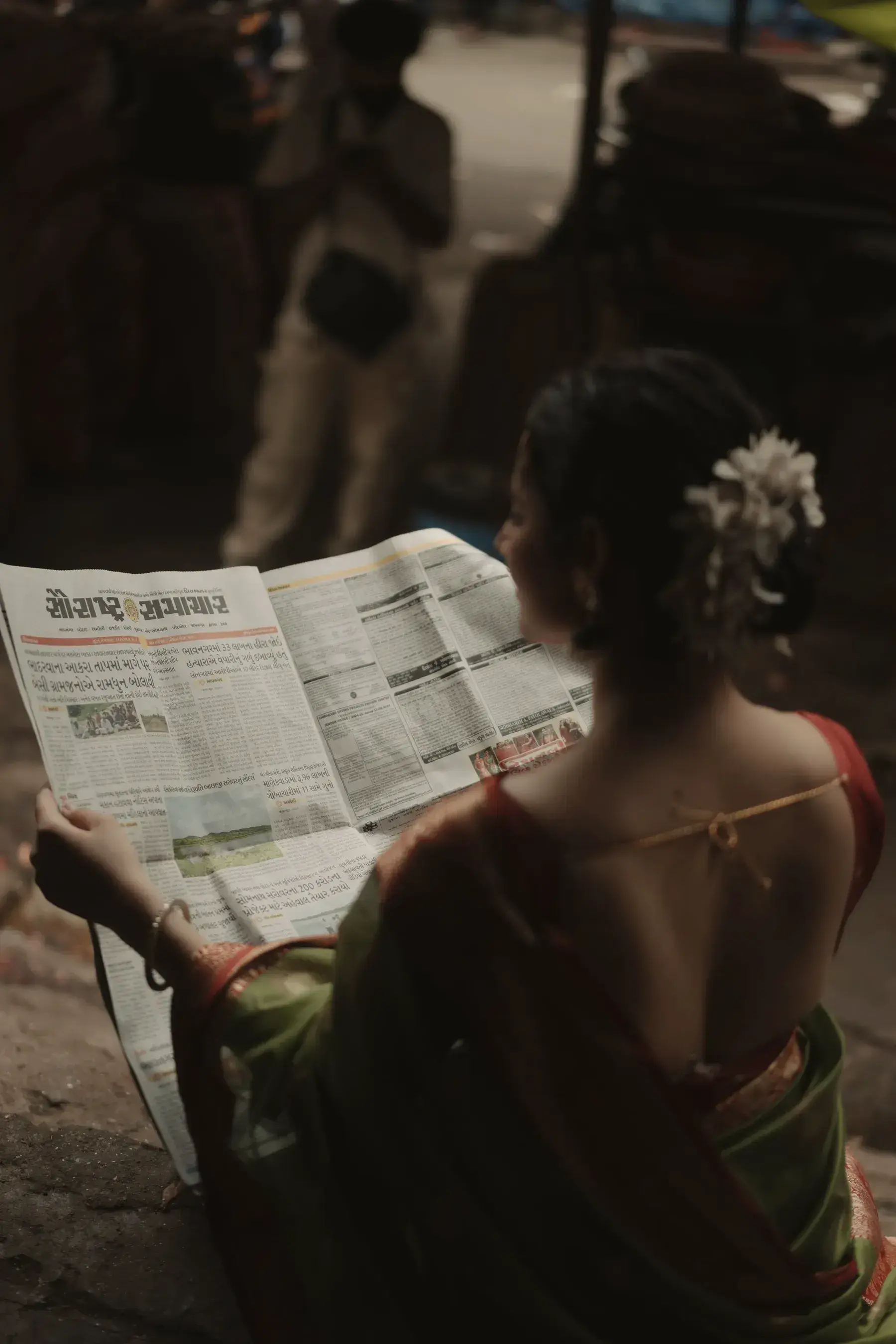 A woman in a saree reading a newspaper, seen from behind, a white flower in her hair