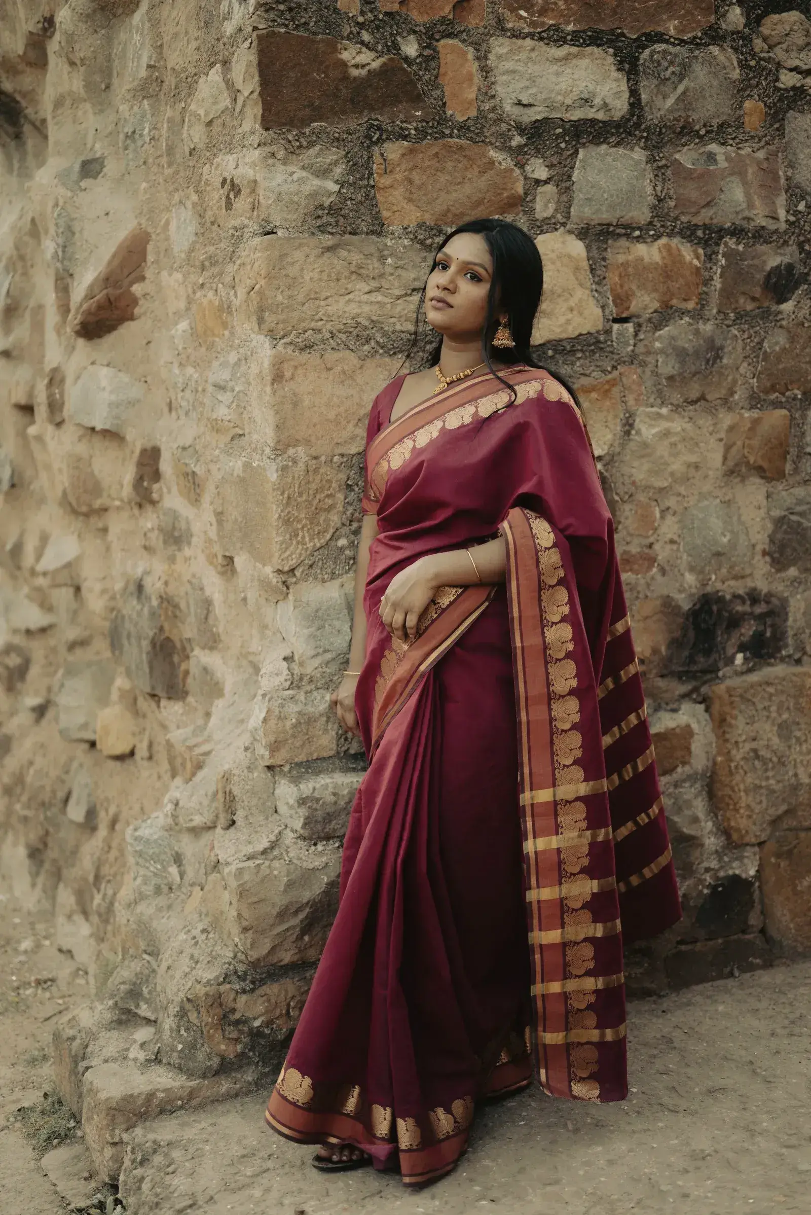 A woman in a maroon tussar saree with gold stripes against a stone wall