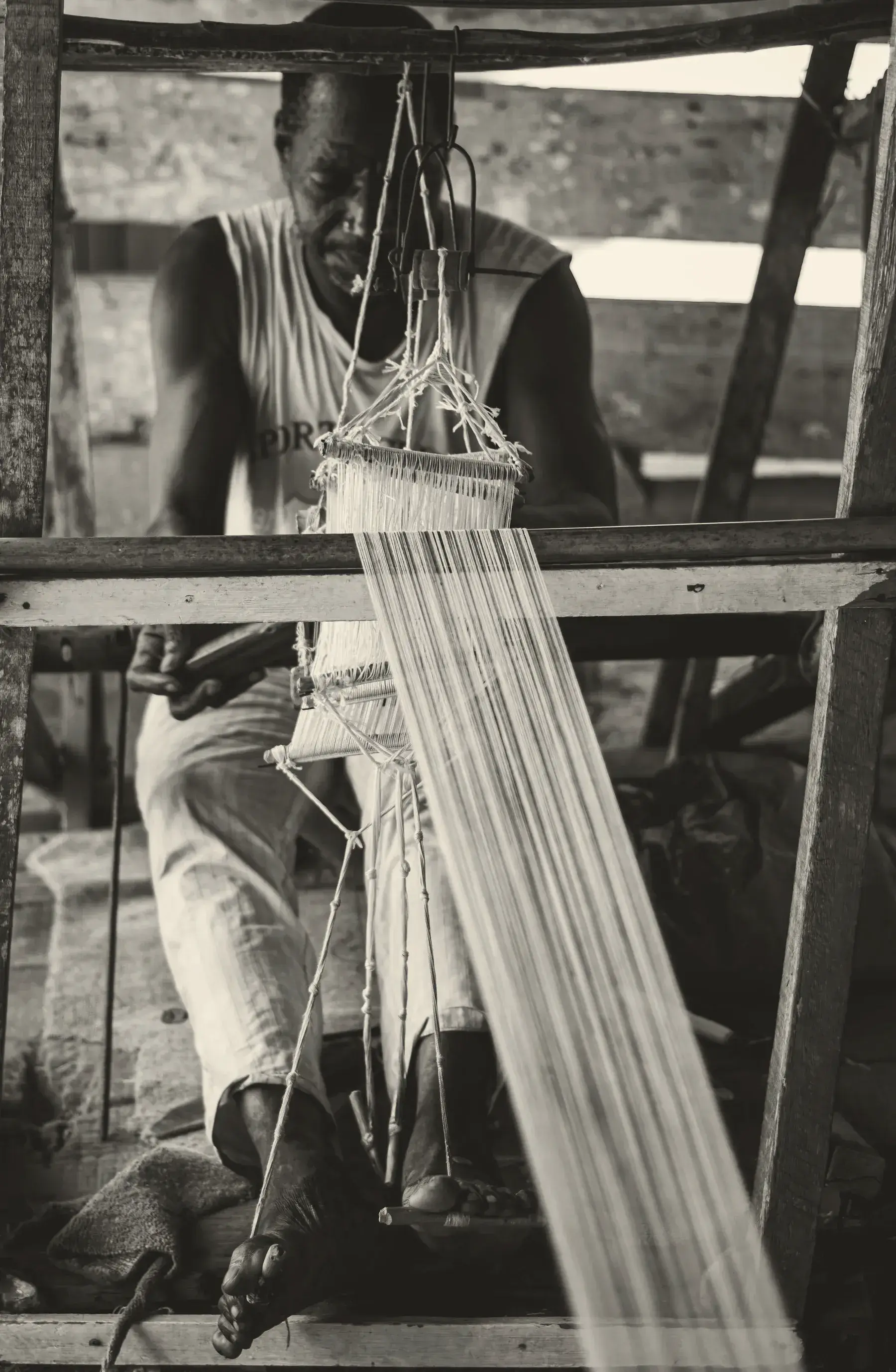 A weaver at a pit loom, the white warp threads running toward the camera, in black and white